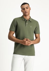 Oliwkowa koszulka polo wykonana z gładkiej bawełny, z klasycznym kołnierzykiem, krótkimi rękawami i małym haftowanym logo na klatce piersiowej.