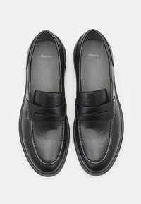 Mocasines de cuero negro con una textura suave, puntera redonda y acentos decorativos cosidos en la parte superior. Plantillas acolchadas visibles en el interior.