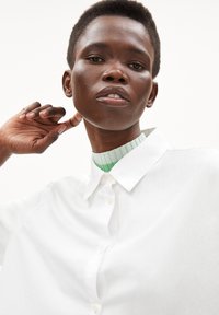 White button-up shirt with a smooth texture, featuring a pointed collar over a green ribbed sweater. Close-up view of facial features.