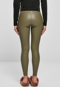 Persona che indossa leggings in finta pelle verde oliva, maglietta nera a maniche lunghe e pantofole marroni pelose, in piedi contro uno sfondo chiaro e uniforme.