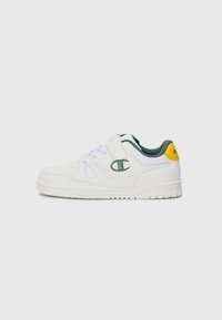 Chaussure de sport blanche avec des accents verts et jaunes ; comporte une tige texturée, une sangle ajustable et une semelle en caoutchouc avec un motif de crampons.