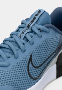 Blue mesh athletic shoe with black accents, textured surface, rounded toe, and contrasting grey midsole. Features laces and a logo tag.