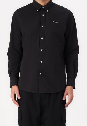 Mann trägt ein schwarzes, langärmeliges Hemd mit Knöpfen und dem "Barbour"-Logo auf der Brust, kombiniert mit schwarzen Cargohosen.