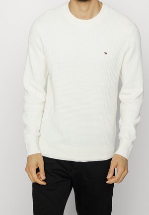 Mężczyzna ubrany w białą teksturowaną bluzę z długim rękawem z małym logo na piersi oraz czarne spodnie, stojący z rękami swobodnie opuszczonymi wzdłuż ciała.