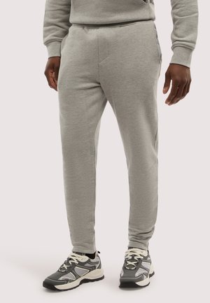 Mann trägt hellgraue Jogginghose und graue und weiße Sportschuhe, steht vor einem schlichten hellen Hintergrund.