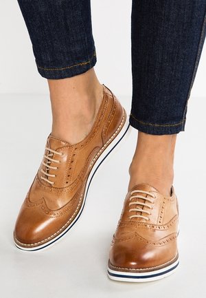 Brune læderbrogues med dekorative perforeringer, rundet tå og en hvid stribe på sålen. Har matchende snørebånd og syninger.