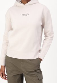 Sudadera de algodón color beige claro con bolsillo frontal, capucha con cordones y logotipo "CALVIN KLEIN NEW YORK" bordado en gris.