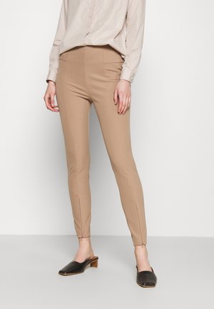 Frau trägt beige, eng anliegende Hose, hellbeige Bluse und schwarze flache Slipper, steht vor einem einfarbigen hellen Hintergrund.