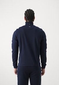 Marineblauwe sporttrui met een hoge kraag, lange mouwen en geribbelde manchetten, met het Fila-logo op de achterkant. Soepele textuur.