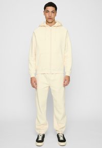 Kremfarget hettegenser med glidelås foran, elastiske mansjetter og matchende joggebukser. Svarte og hvite sneakers fullfører antrekket. Mykt, teksturert stoff.