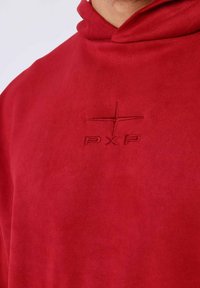Sudadera con capucha roja hecha de tela suave, con un logo bordado, ligera textura y un corte holgado.