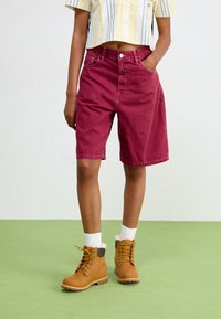 Shorts in denim borgogna con una vestibilità morbida, tasche frontali e chiusura a bottone, abbinati a stivali in tan con lacci e colletto morbido.