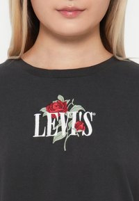 Juodas medvilninis marškinėlis su baltu "LEVI'S" logotipu, virš kurio išausti raudoni rožės ir žali lapai. Apvalaus iškirpimo dizainas.