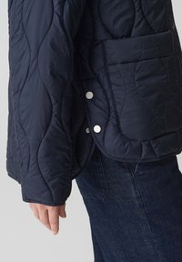 Giacca trapuntata color navy con tasca laterale, bottoni a pressione argentati e una texture isolante, abbinata a jeans in denim scuro.