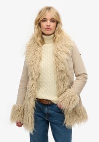 Beige jasje met een donzige beige bontkraag en manchetten, gecombineerd met een crèmekleurige kabelgebreide trui en denim jeans.