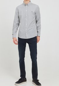 Camicetta grigia con un sottile motivo a righe verticali, abbinata a jeans scuri e sneakers nere; presenta un colletto classico e un logo sul petto.
