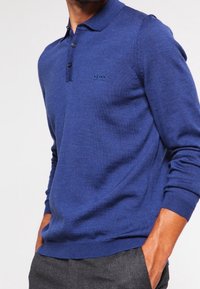 Polo en tricot bleu avec une patte de boutonnage à trois boutons, des poignets côtelés et un petit logo sur la poitrine gauche. Texture douce et design ajusté.