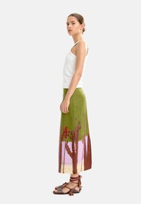 Zielona spódnica do kolan z kolorowym nadrukiem przedstawiającym drzewa i krajobraz; modelka nosi białą bluzkę na ramiączkach i sandały na cienkim obcasie.