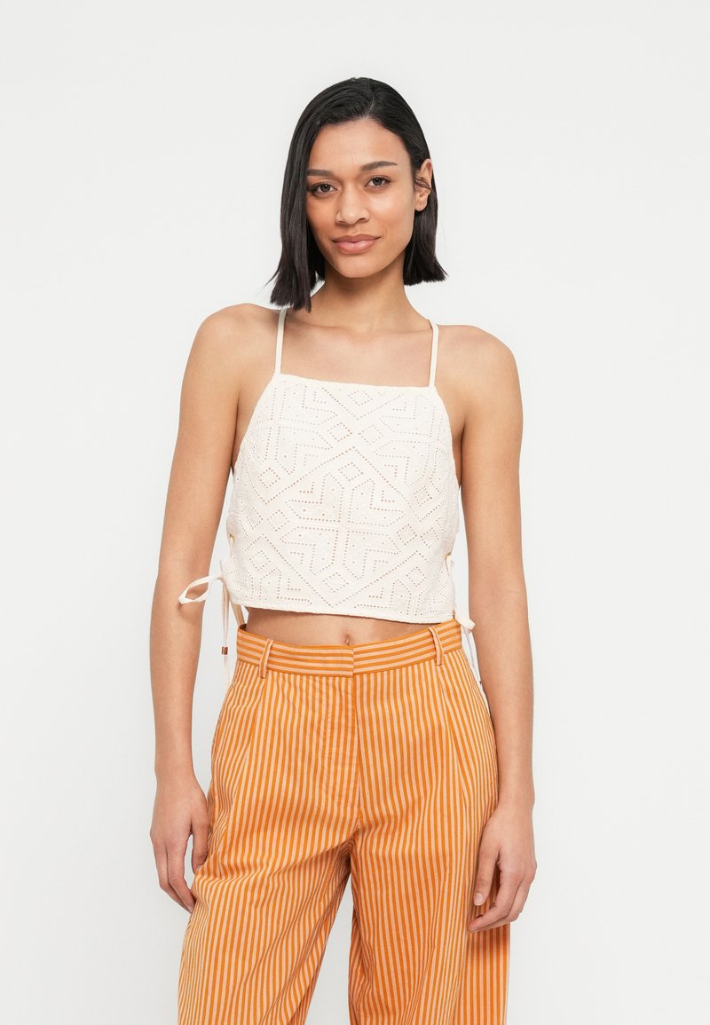 Blouse crème raccourcie avec motifs géométriques ajourés, côtés ajustables avec lien, associée à un pantalon taille haute à rayures orange et blanches.