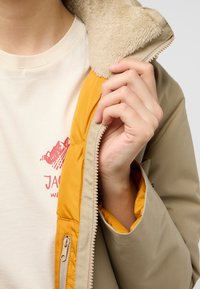 Veste beige à fermeture éclair avec un col beige doux et une doublure orange vif, présentant une main à l'intérieur de la veste et un logo visible sur une chemise crème.