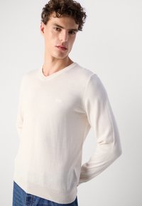 Maglione a V in maglia avorio, caratterizzato da maniche lunghe e delicati dettagli a coste sui polsini e sull'orlo. Logo ricamato sul petto.
