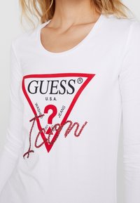 Camicia bianca a maniche lunghe con un logo triangolare rosso e il testo "GUESS" e "U.S.A.", con elementi in strass che formano la parola "toan."