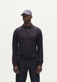 Tengerészkék hosszú ujjú pólóing, galléros nyakkal és gombos díszítéssel, sötét farmernadrággal és egy hozzá illő világoskék sapkával párosítva.