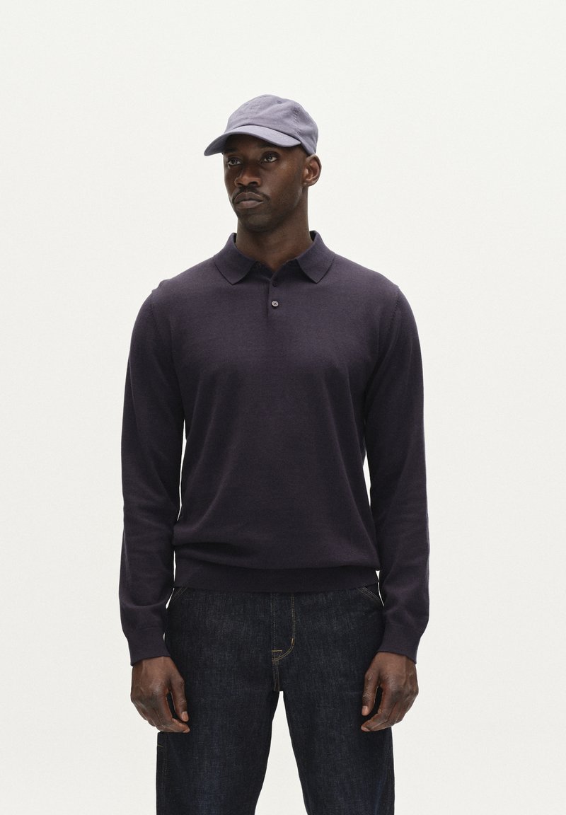 Tengerészkék hosszú ujjú pólóing, galléros nyakkal és gombos díszítéssel, sötét farmernadrággal és egy hozzá illő világoskék sapkával párosítva.