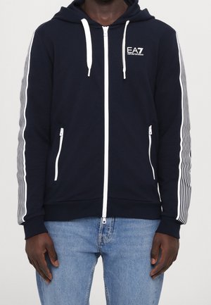 Homme portant un sweat à capuche zippé bleu marine avec logo blanc EA7 Emporio Armani et manches à rayures, assorti à un jean bleu clair.
