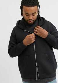 Felpa nera con zip e tasca frontale, caratterizzata da una texture liscia, zip argentata e cappuccio con coulisse. Indossata sopra una maglietta nera.