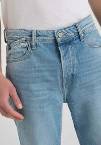 Jeans de mezclilla azul claro abrochados con un botón, usados con una camisa blanca metida por dentro, mostrando la cintura frontal y la mano izquierda.