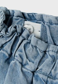 Ljusblå denimgabardin med samlade detaljer, försedda med en vit knapp och en texturerad midjeband. Sömmen är synlig och välgjord.