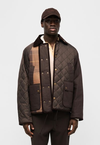 Άνδρας που φοράει καφέ καπιτονέ μπουφάν με λεπτομέρεια καρό, κουμπιά τύπου σναπ, μεγάλες τσέπες και καπέλο σε καφέ απόχρωση, μπροστά σε απλό φόντο.