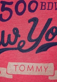 Camiseta de algodón roja con texto bordado en azul marino, que dice "Te Queremos" en cursiva y "TOMMY" en un parche rectangular beige.