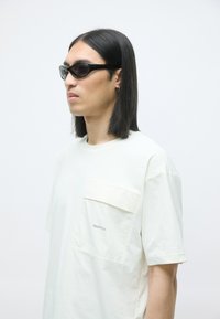 Lys beige oversized t-skjorte med brystlomme, laget av myk bomull. Har korte ermer og diskret merkevare ved kanten. Svart ovalt solbriller.