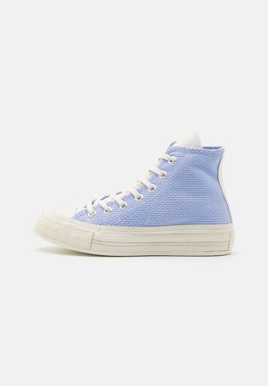 Sneakers montantes avec dessus en tissu texturé bleu clair, semelle en caoutchouc blanche, bout renforcé et lacets blancs sur fond blanc.