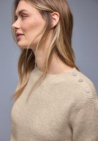 Femme aux longs cheveux blonds portant un pull en tricot beige avec trois boutons sur l'épaule, sur un fond gris uni.