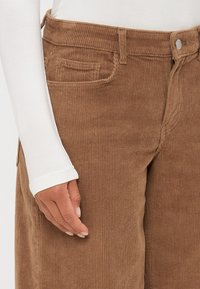Bruine corduroy broek met geribbelde textuur, voorzien van steekzakken, een knoopsluiting en een hoge taille. Een witte blouse met lange mouwen is gedeeltelijk zichtbaar.