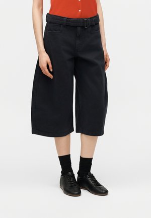 Άτομο που φοράει μαύρο φαρδύ cropped παντελόνι με ζώνη, μαύρες κάλτσες, μαύρα δετά παπούτσια και πορτοκαλί αμάνικη μπλούζα.