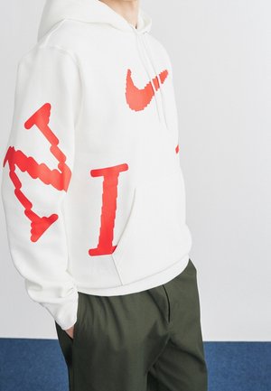 Osoba nosící bílou mikinu s pixelovaným červeným logem Nike a písmeny, doplněnou tmavě zelenými kalhotami, ruce v kapsách.