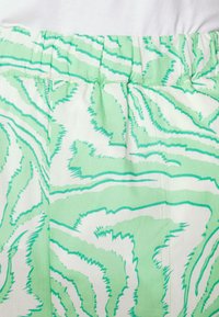 Tecido patterned verde e branco com linhas onduladas, apresentando um cós elástico e uma textura suave. O design enfatiza fluidez e movimento.