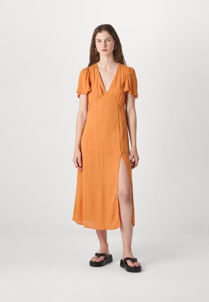 Orange midi klänning med djup V-ringning, korta fladderärmar, sidostygn och lätt tyg; matchad med svarta platta sandaler.