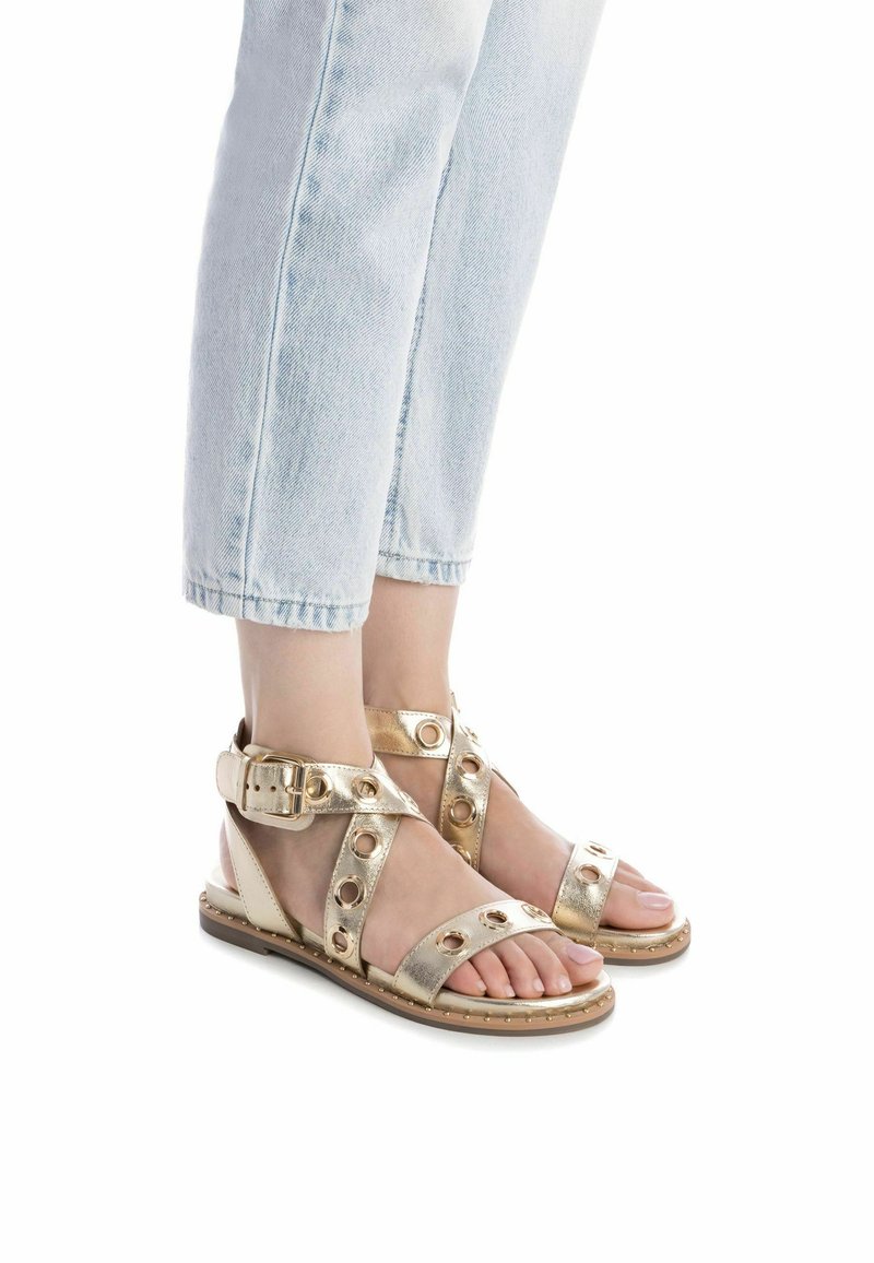 Sandalias metálicas doradas con correas anchas que presentan cortes circulares, una correa en el tobillo con hebilla y una suela plana, combinadas con jeans recortados de color azul claro.