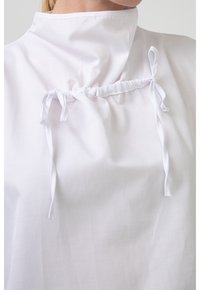 Witte blouse met een hoge kraag, voorzien van een geplooide voorkant met twee witte strikjes voor aanpassing. Glad stof met een lichte glans.
