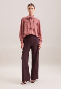 Blusa rosa con fiocco e bottoni, abbinata a pantaloni borgogna a vita alta e gamba larga e sandali neri con tacco. Texture morbida.