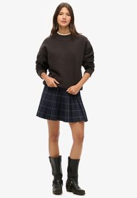 Sweat-shirt noir à épaules tombantes, jupe plissée à carreaux bleu marine, et bottes noires montantes avec détail de boucle. Look simple et décontracté en couches.