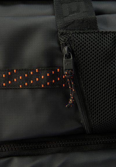 Gros plan sur un tissu noir avec une sangle orange à pois, une fermeture éclair avec tirette, et un détail de poche en maille sur un sac.