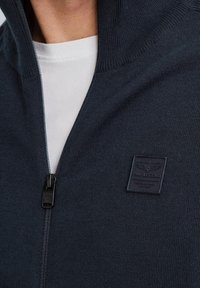 Navy zip-up sweater made of knit fabric; features a high collar, a lightweight zipper, and a textured patch label on the chest.