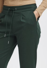 Tmavě zelené joggingové kalhoty s elastickým pasem, stahovací šňůrkou, bočními kapsami a měkkou, hladkou texturou látky.
