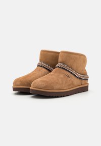 Brune suede ankelstøvler med et dekorativt mønstret bånd rundt om skaftet, en brun gummisål og prægede logo på siden.